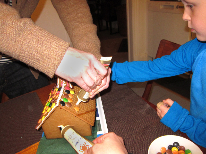 På bilden syns två händer som spritsar glasyr på taket av ett pepparkakshus. I kanten av bilden syns ett barn i blå tröja som deltar i dekoreringen. Taket är dekorerat med färgglada godisar och vit glasyr.