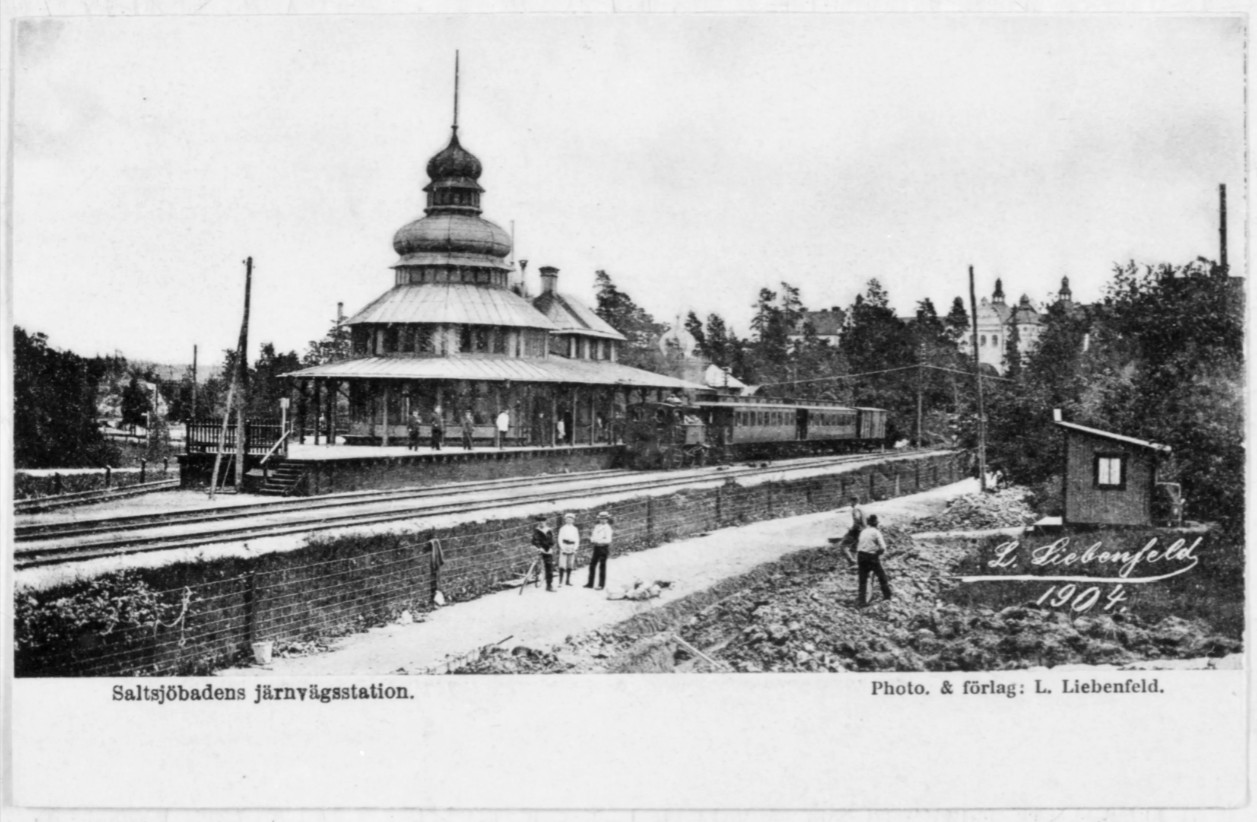 En byggnad ligger precis bredvid järnvägsstationen, några människor står vid huset.