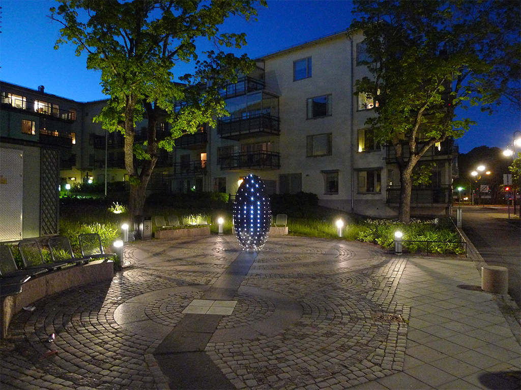 Lysande skulptur i blått i ett bostadsområde med träd och stenläggning.