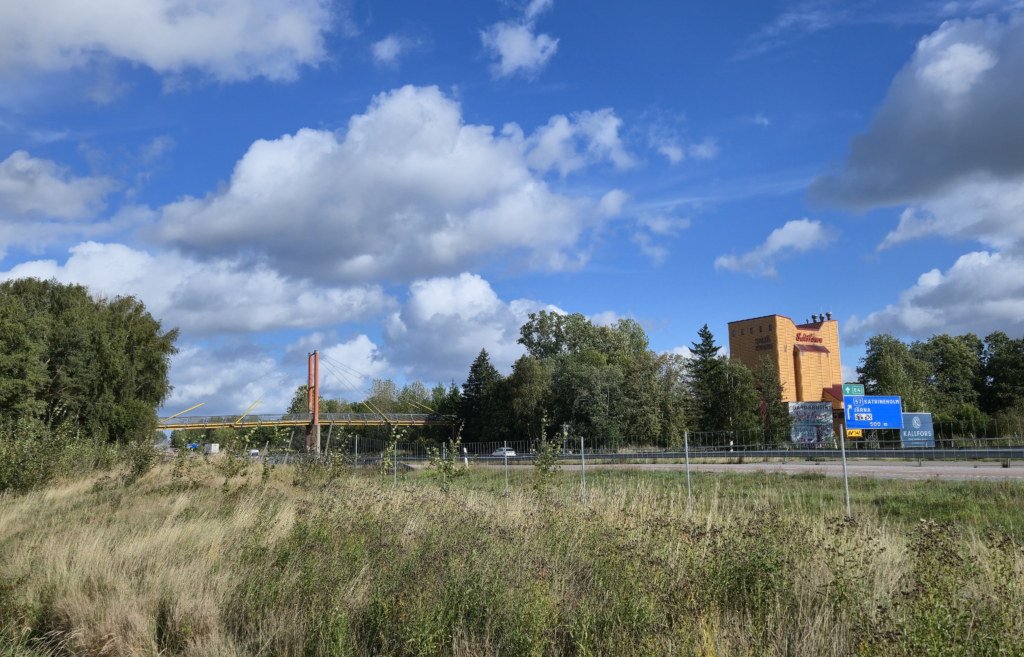 Åkermark, en motorväg och ett gult hus i bakgrunden. Över vägen går en färgglad bro.