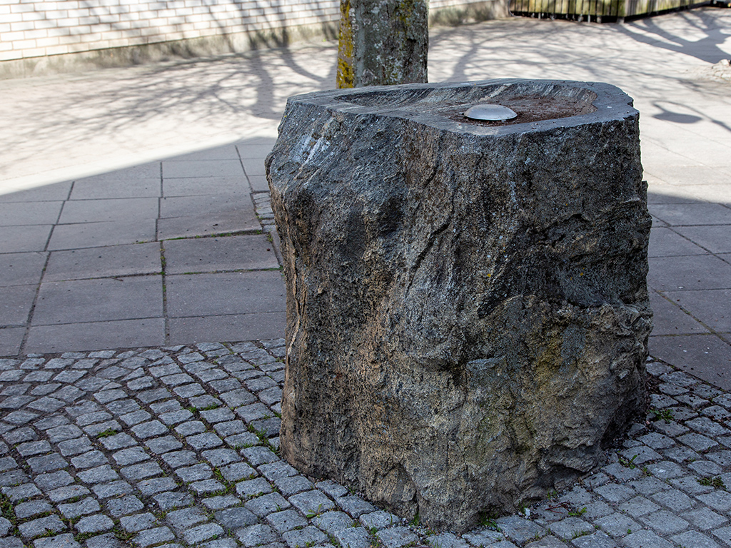 Oregelbundet formad sten med en slipad kant och en fördjupning i mitten. Står på en stenläggning med smågatsten.