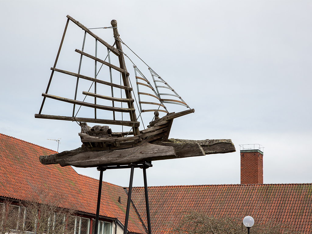 Träbåt med segel mot en grå himmel framför hus med tegeltak.