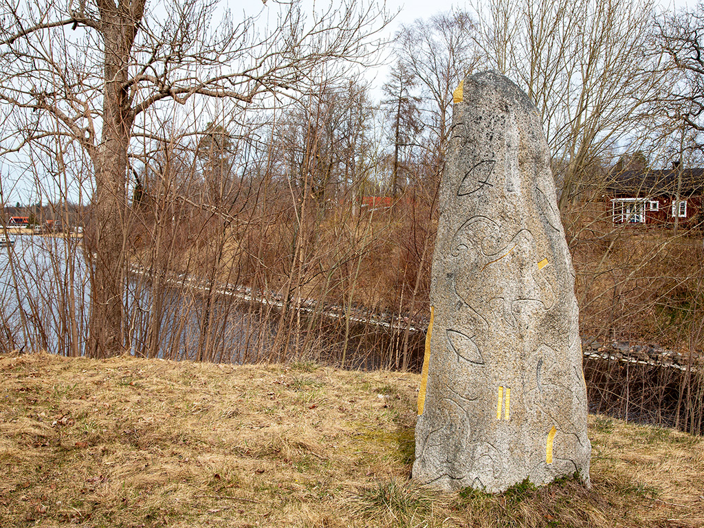 En hög sten med ristningar och fält av guld på vissa ställen. Står i gräset vid en kanal.