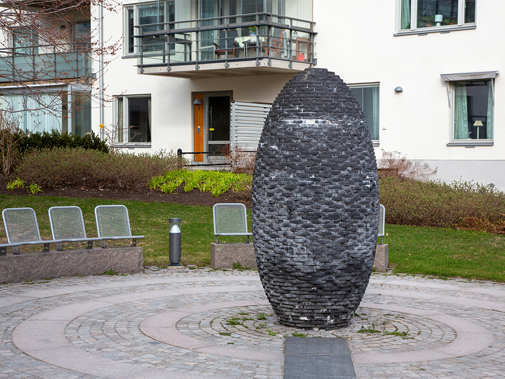 Cylinderformad skulptur i mörk murad sten på rundel av gatsten framför ett flerbostadshus.