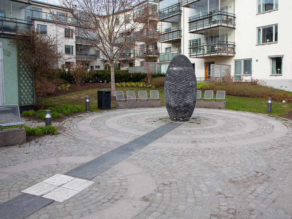 Cylinderformad skulptur i mörk murad sten på rundel av gatsten framför ett flerbostadshus.
